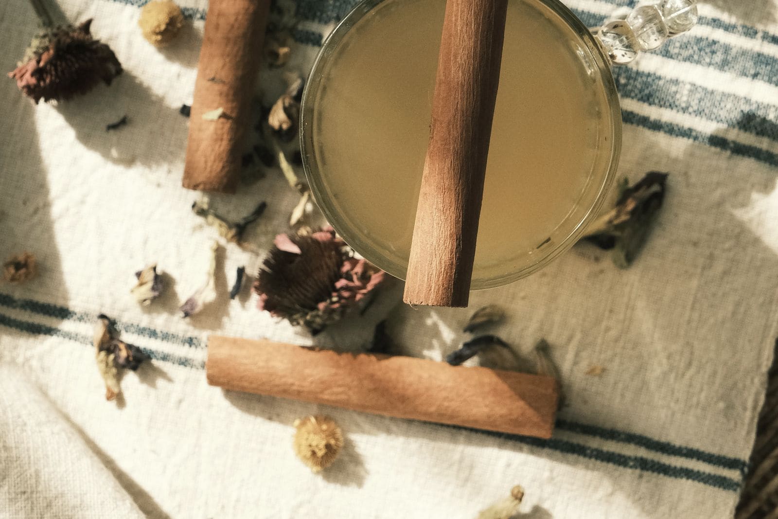 A glass of herbal tea with cinnamon sticks and dried flowers on a striped linen cloth
